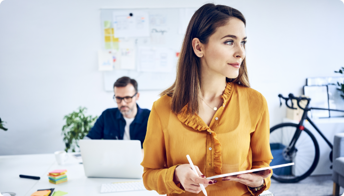 A woman with a clipboard with a man on a laptop in the background.